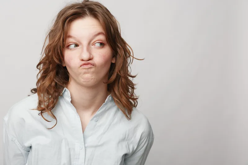 Young woman with curly hair wearing white shirt makes funny face looking sideways. Studio portrait on light background. Concept of humor, expression, and casual lifestyle.