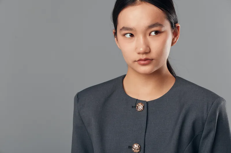 Young Asian woman wearing a dark gray blazer with decorative buttons looking to the side on gray background. Portrait, confident expression, professional style, studio lighting, isolated.