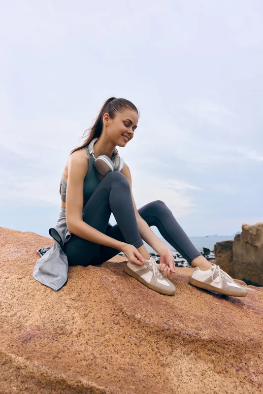 Fit young woman tying her sneakers on a rocky beach in activewear, showcasing a healthy lifestyle and outdoor exercise