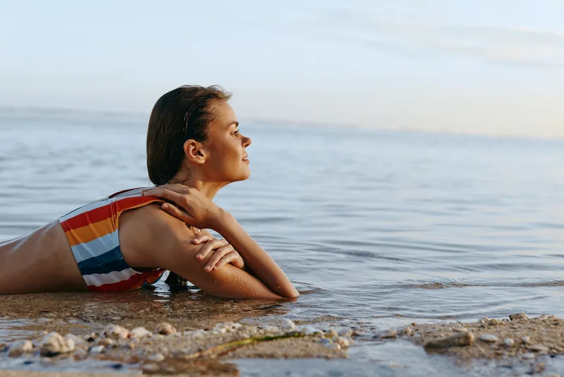 Young woman in striped swimsuit lies relaxed on the beach shore with clear calm sea water around, enjoying peaceful sunny day and fresh air by the ocean.