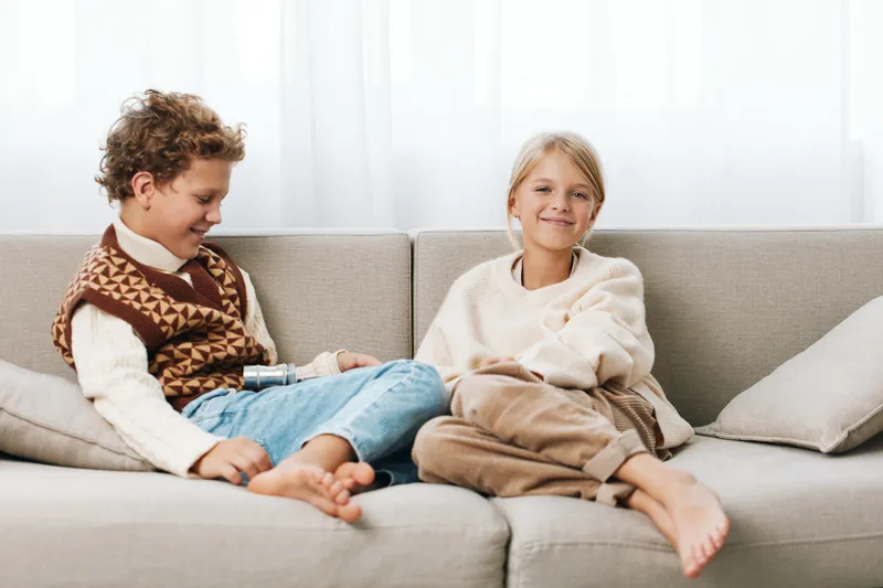 Two children sit on a light grey sofa, smiling and relaxed, wearing cozy sweaters. The scene captures warm friendship, quiet comfort, and everyday home harmony.
