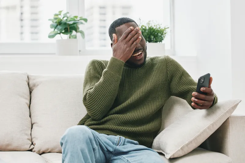 A young Black man in a green sweater expresses frustration while looking at his smartphone in a modern living room filled with plants