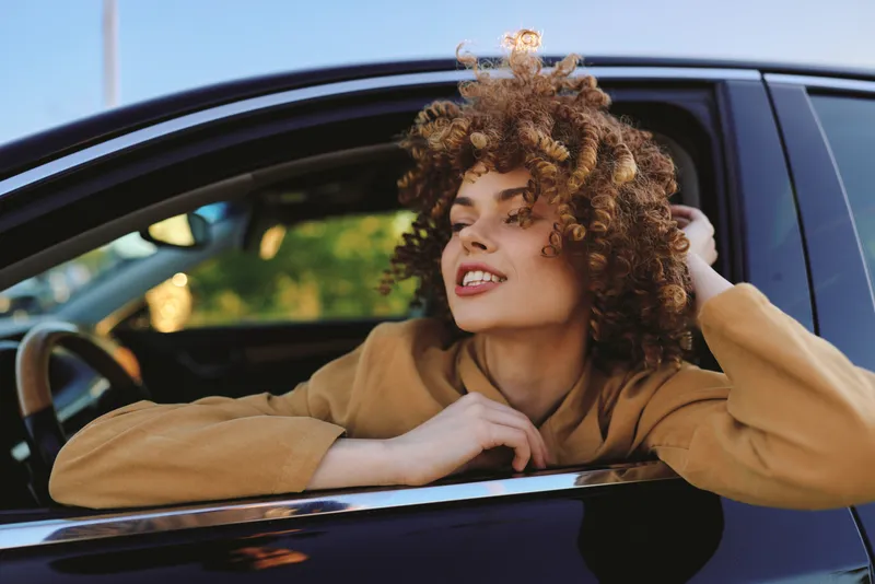 Smiling woman leaning from a car window, casual street style and curly hair. Warm retro mood with natural light and film color evokes relaxed lifestyle and candid confidence.