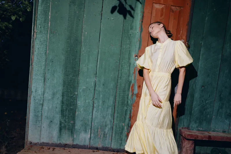 Fashionable woman in a flowing yellow dress leaning against a green wooden wall in an outdoor setting, captured in moody light for a cinematic portrait with soft shadows and elegance.