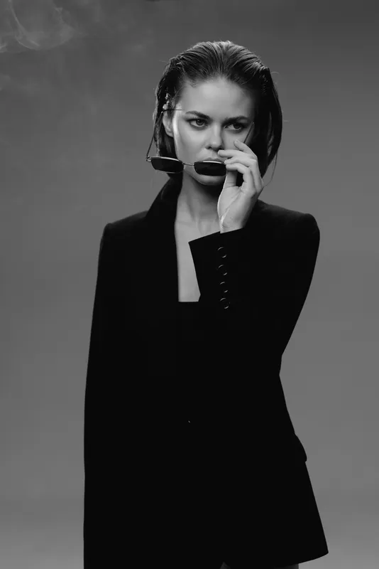 Black and white portrait of a serious woman in a dark blazer holding sunglasses with a moody expression