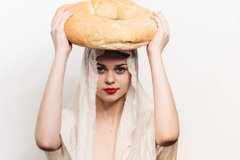 Счастливая женщина с тканью на голове и буханкой хлеба, мучным изделием, на светлом фоне; место для копирования