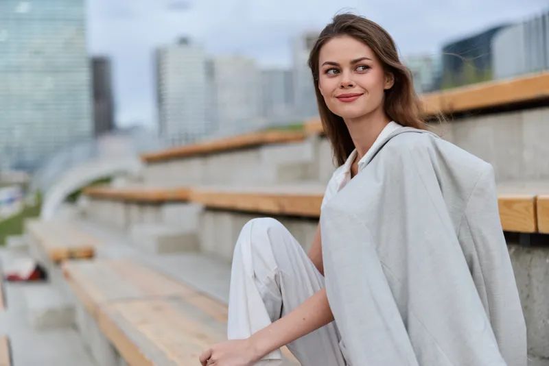 young woman wearing light gray coat sitting on urban bench with modern cityscape background confident and stylish outdoor fashion portrait