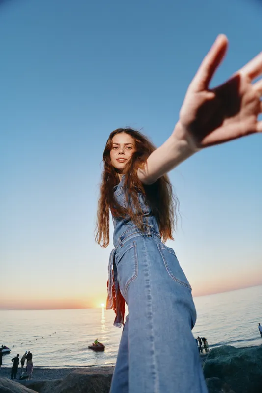 lifestyle woman in modern boho-western denim street style reaching out with film color tones at sunset near the sea with natural light and relaxed mood