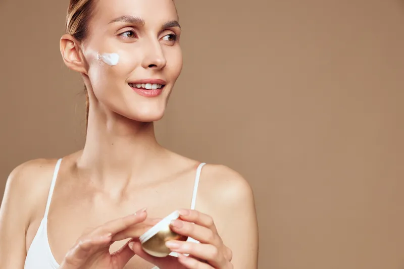 A young woman in a white tank top applies face cream on her cheek with a smile. The neutral background highlights skincare, beauty, and healthy lifestyle concepts.