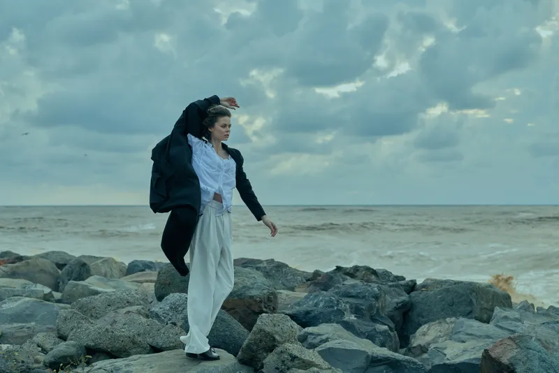 Sea shore scene with a woman on rocks, wind-swept coat and flowing dress, capturing a dramatic outdoor fashion moment by the ocean and rugged shoreline