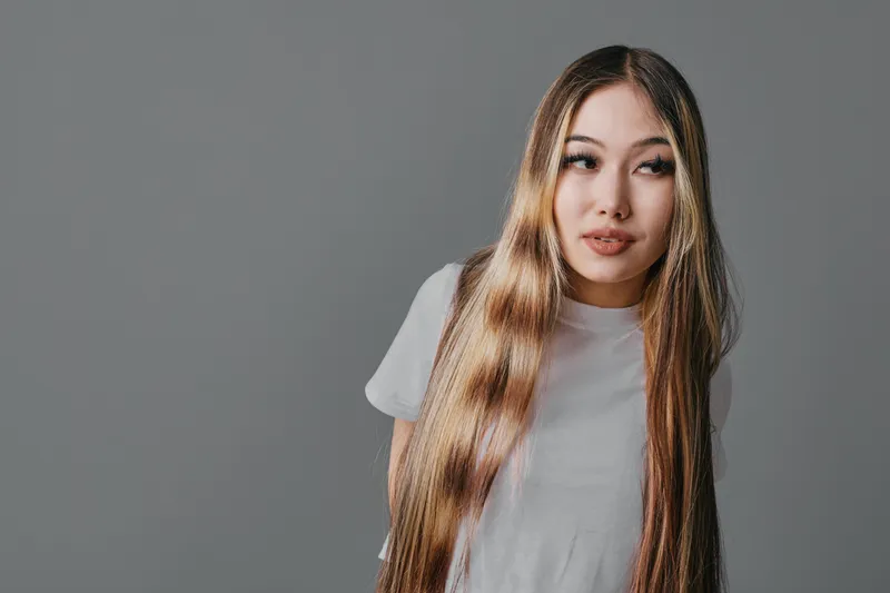 Young woman with long straight hair and casual white t-shirt posing against gray studio background looking sideways with thoughtful expression and natural makeup.
