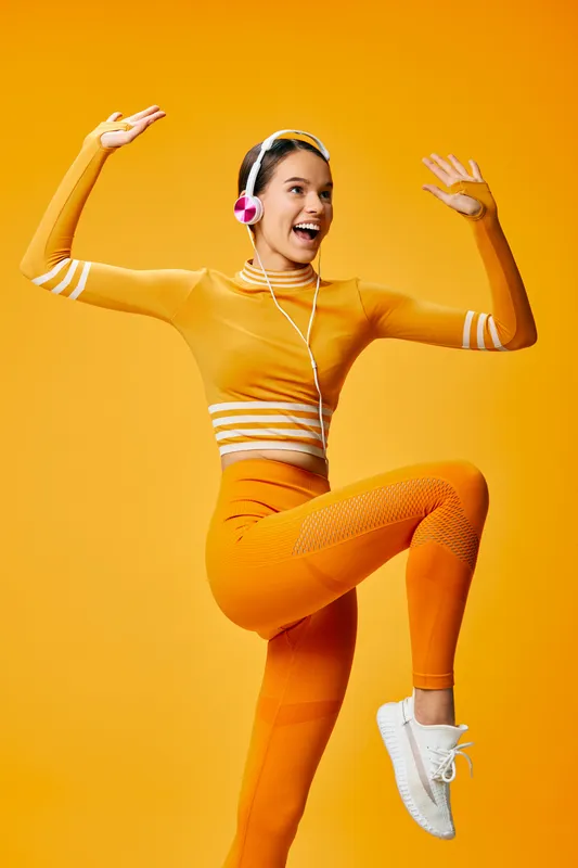 Energetic woman in orange fitness outfit dancing and jumping with headphones against a vivid orange studio backdrop, capturing movement, rhythm, and active lifestyle.