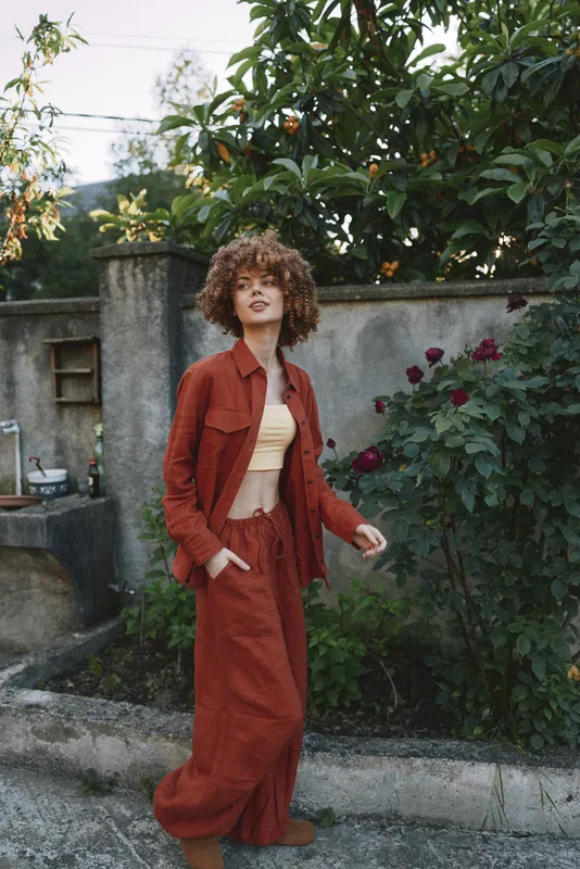 Curly-haired woman in rust outfit standing in a garden, casual lifestyle shot. Natural light highlights warm tones and greenery around. Focus on fashion and confidence