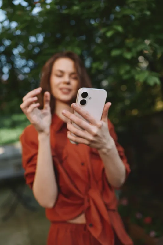 A woman in a rust outfit takes a casual outdoor selfie, smiling at her phone, capturing a relaxed lifestyle moment in natural light and greenery, ideal for contemporary fashion and everyday
