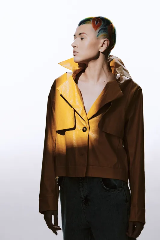 Young woman with colorful stylish short hair wearing brown leather jacket and denim, posing against a bright white background with dramatic lighting and shadows.