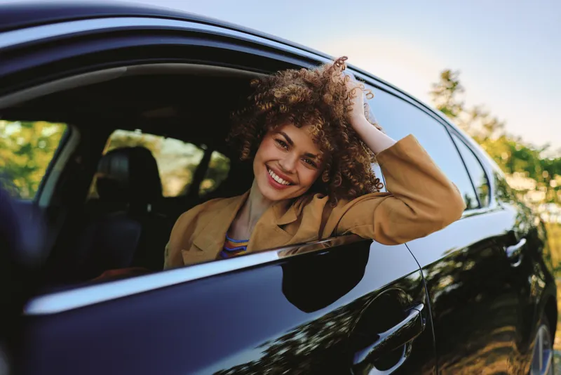 A smiling woman leans out of a sleek car window, wearing a casual jacket, as warm sunlight floods the scene. An upbeat lifestyle moment captured outdoors with freedom.