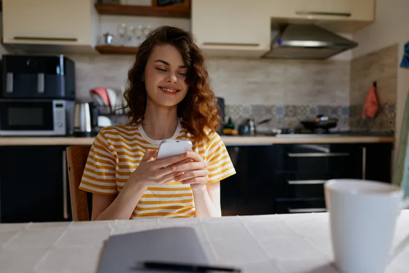 Woman relaxing at home in a bright kitchen, smiling while looking at her phone, casual lifestyle moment, warm light, modern interior, table with mug and cups, intimate everyday scene