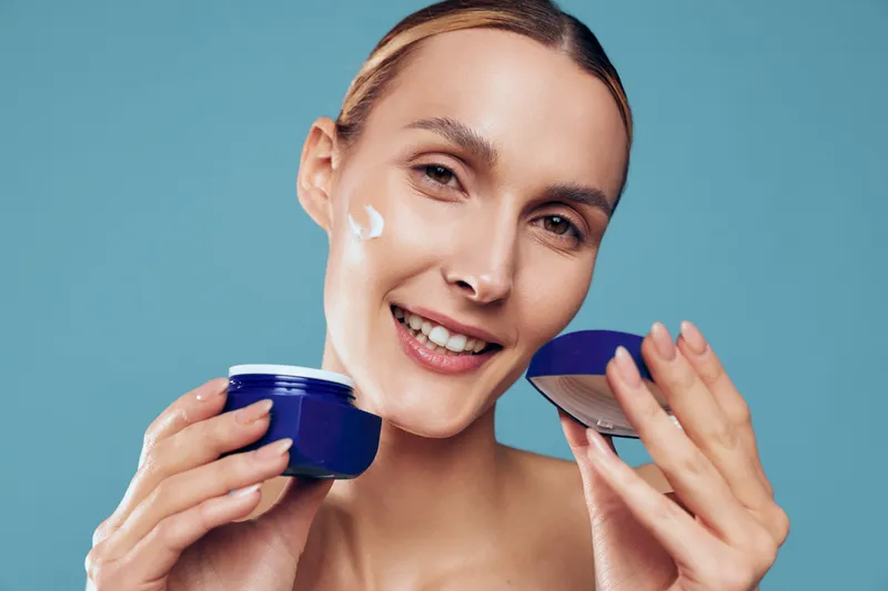 Smiling woman applying white face cream on cheek holding open blue jar against blue background. Concept of skincare, beauty, self-care, moisturizing, and healthy skin routine.