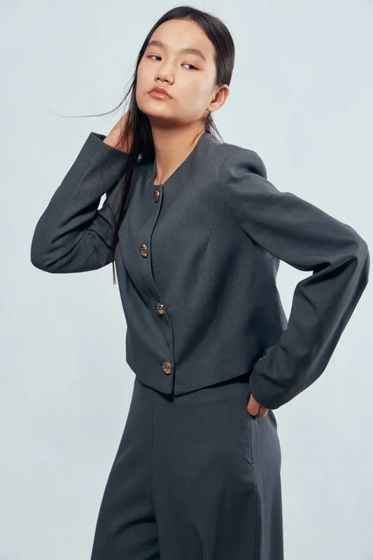 Woman posing in gray stylish suit with buttons, long sleeves and high waist pants on light background. Fashion model with straight hair standing with hand behind head and serious look.
