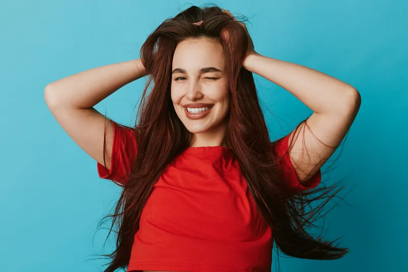 Young woman with long hair and red shirt smiling with eyes closed and hands in hair on blue background. Happy female portrait with natural expression and casual style.