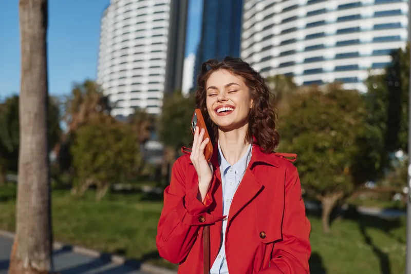 Smiling woman in a red coat talks on a phone outdoors in a sunny park, with modern buildings in the background, casual urban portrait.