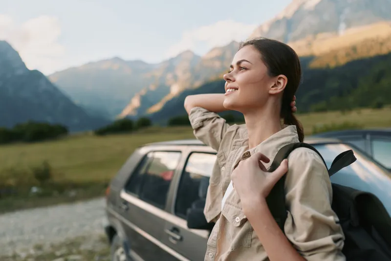 Young woman traveler stands near car in mountains enjoys fresh air and nature, holds backpack strap, smiles with relaxed lifestyle and positive mood outdoors.