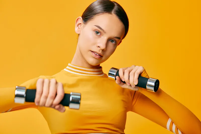 Female fitness enthusiast in a bright yellow studio holds dumbbells with focused expression, showcasing a dynamic workout moment, strong arms, athletic posture, and healthy lifestyle through strength