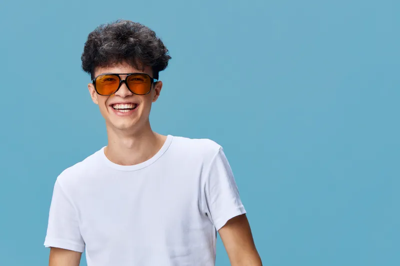 smiling man wearing orange sunglasses and white t-shirt, joyful young adult with curly hair against blue background expressing happiness and style in casual attire
