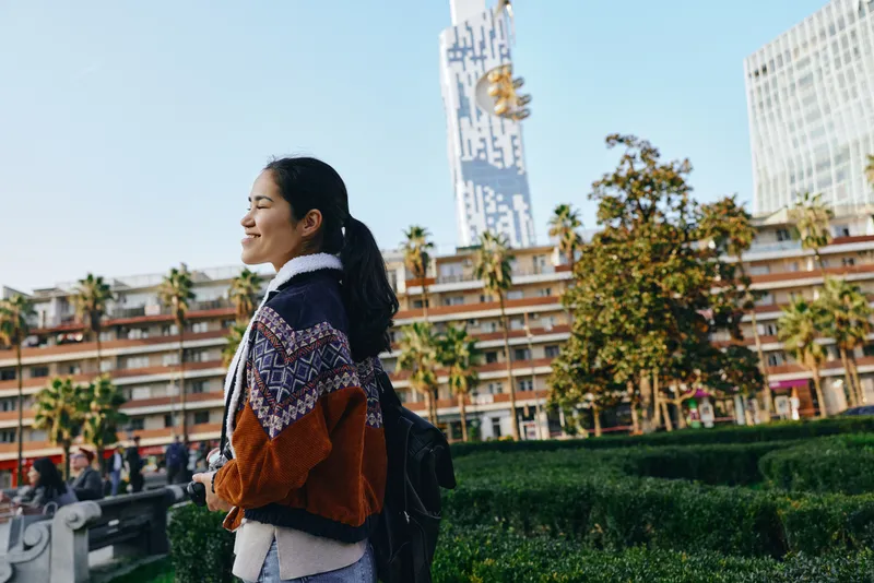 Outdoor urban scene with a woman carrying a backpack, strolling through a sunny city park as modern buildings rise in the distance, casual fashion and exploratory mood.