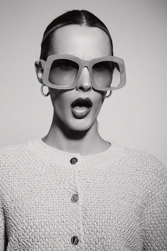 Young Caucasian woman with light skin wearing oversized sunglasses and knitted sweater shows surprised expression in black and white portrait. Concept for fashion and emotions.