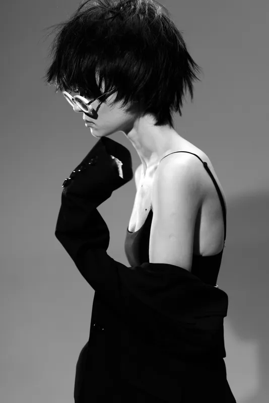 Black and white portrait of a young woman with short hair wearing glasses and a black top, expressing thoughtful elegance and style.
