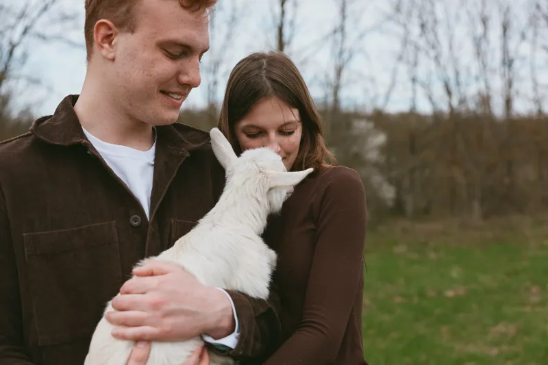 Cheerful couple outdoors, holding a white goat kid, warm embrace, soft natural light, relaxed mood, rustic countryside setting, wholesome lifestyle moment, intimate joyful connection