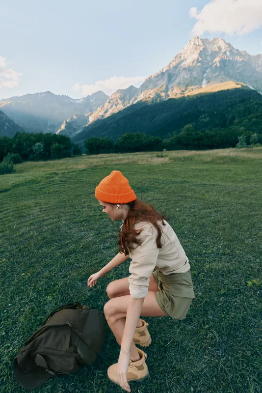 Young woman wearing an orange beanie kneels in a meadow with a backpack against a mountainous backdrop. She studies the ground in a light sweater and shorts, creating a candid outdoor moment.
