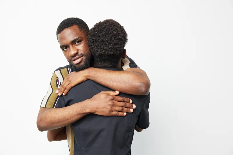 Two black men sharing a warm embrace, expressing friendship and solidarity. The image features a minimalist background that enhances their connection.