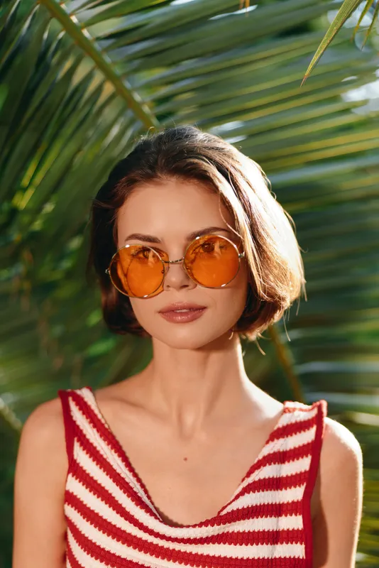 A stylish woman wearing round orange sunglasses stands outdoors among tropical greenery. She wears a red and white striped top and smiles, creating a bright, sunny mood with a chic, contemporary vibe.