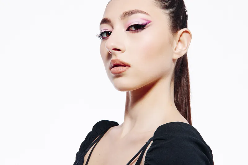 Young woman with creative purple eyeliner and perfectly styled hairstyle poses confidently in black outfit on white background. Concept for beauty, fashion, invention, and makeup art.