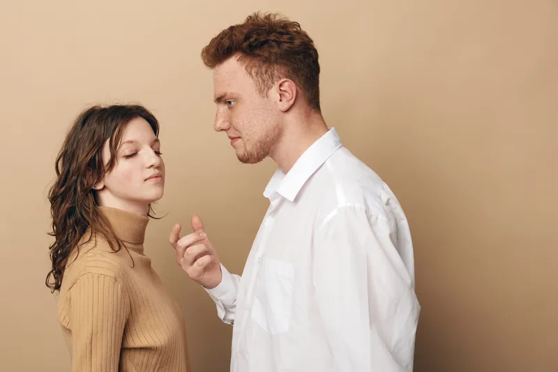 Young man and woman stand close facing each other on beige background. The woman wears a brown turtleneck and the man a white shirt. Concept of communication and relationship tension.