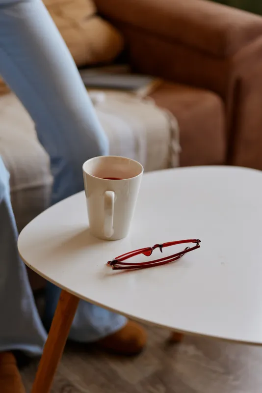 Casual home scene with a white table holding a mug and red glasses, soft living room in the background, relaxed mood, simple everyday moment of morning routine.