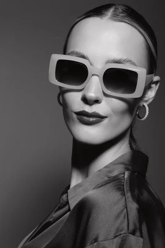 Stylish caucasian woman with pale skin wearing oversized sunglasses and hoop earrings posing in dark clothing studio portrait.