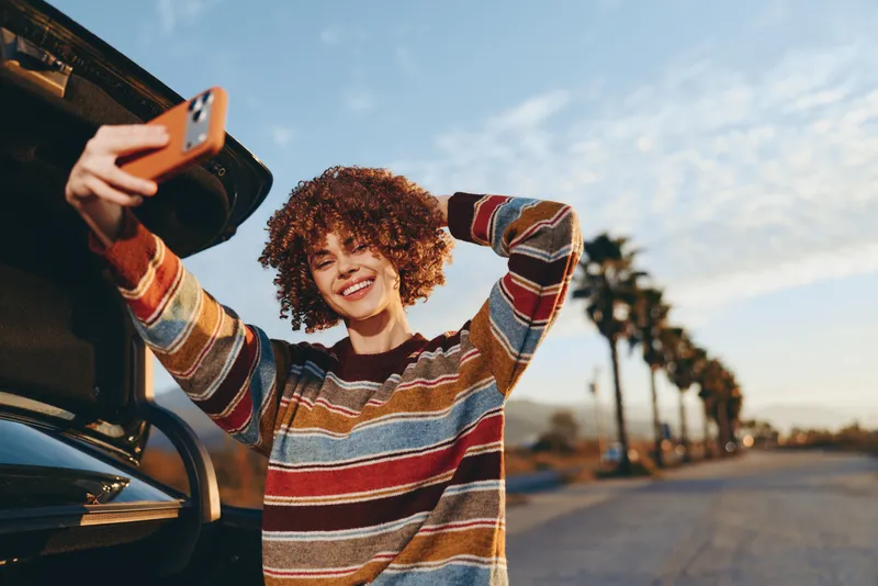 Woman lifestyle scene with a smiling woman holding a smartphone beside a car outdoors at street sunset, curly hair, striped sweater, palm lined road, warm sunlight and relaxed mood