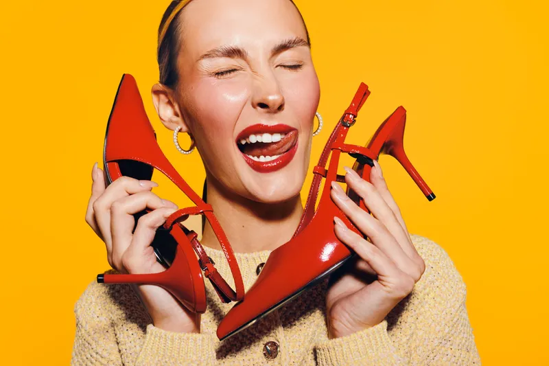 Happy woman with closed eyes holds red high heel shoes near face, wearing beige sweater and gold hoop earrings. Bright yellow background creates vibrant and joyful mood for fashion concept.