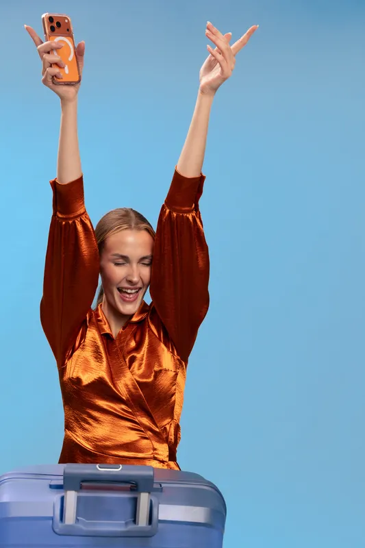 Happy young woman in shiny orange blouse raises arms and smiles while holding smartphone above blue suitcase. Concept of travel excitement, vacation, and joyful journey with luggage.