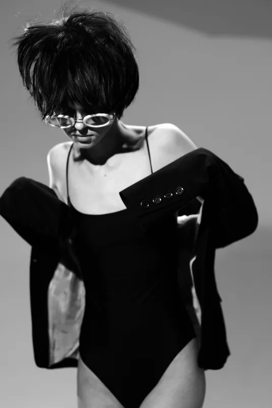 Black and white portrait of a stylish woman wearing sunglasses and a bodysuit with a jacket, showcasing high fashion and modern elegance.