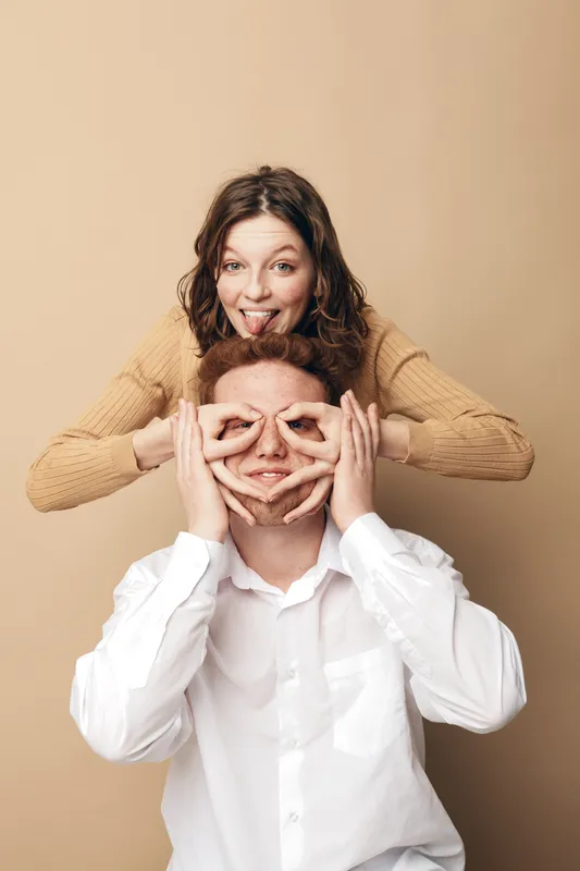 Playful young woman in beige sweater makes funny face behind smiling man in white shirt on beige background. Concept of friendship, fun, happiness, and casual lifestyle together.