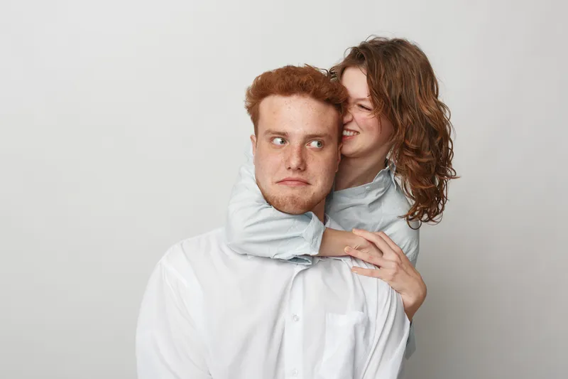 Young woman playfully hugs serious young man from behind, both wearing white shirts, isolated on grey background. Concept of relationship, affection, and playful interaction.