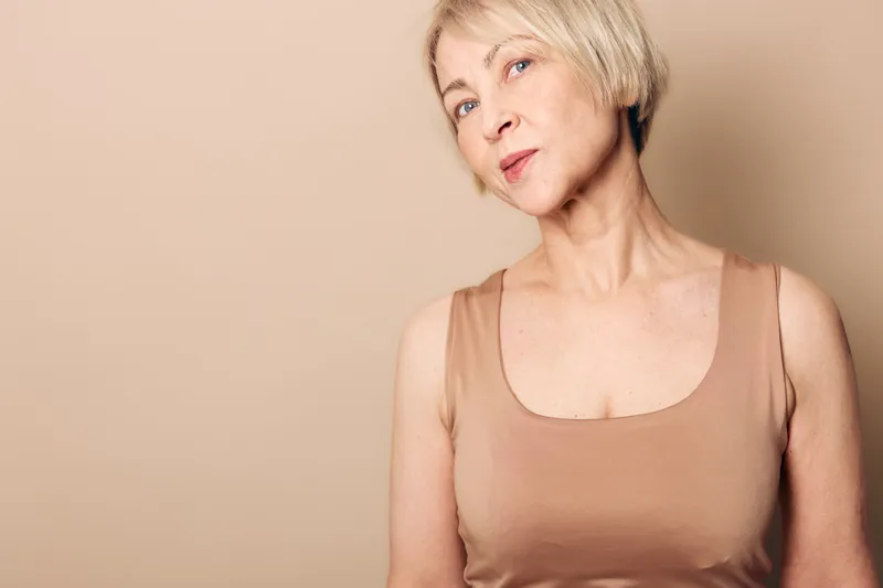 Portrait of a confident mature woman with short blonde hair wearing a beige sleeveless top against a neutral background. Concept of natural beauty, aging, and self-confidence.