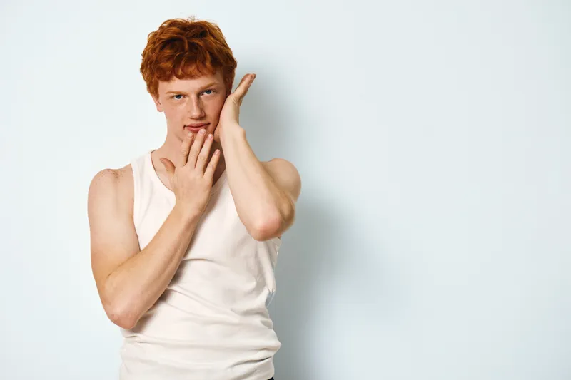 Young man with red hair posing with hands near face wearing a white tank top. Casual fashion, confident expression on light background, portrait of male model.