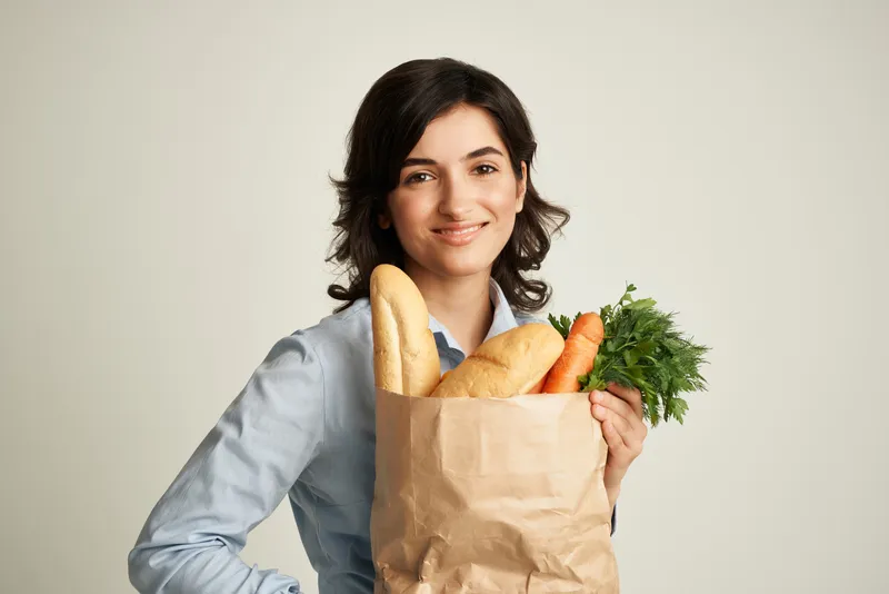 Весёлая брюнетка с пакетом продуктов, полным овощей, совершает покупки здоровой пищи