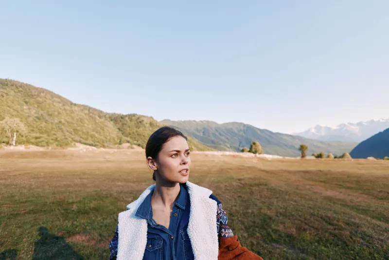 Woman portrait in meadow with mountains landscape, outdoor nature and travel mood. Young female in denim jacket and shearling coat gazing to the side across grassy field.