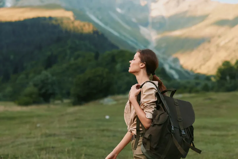 A young woman with a backpack gazes toward towering mountains in a green valley, embodying adventurous exploration and calm in nature.